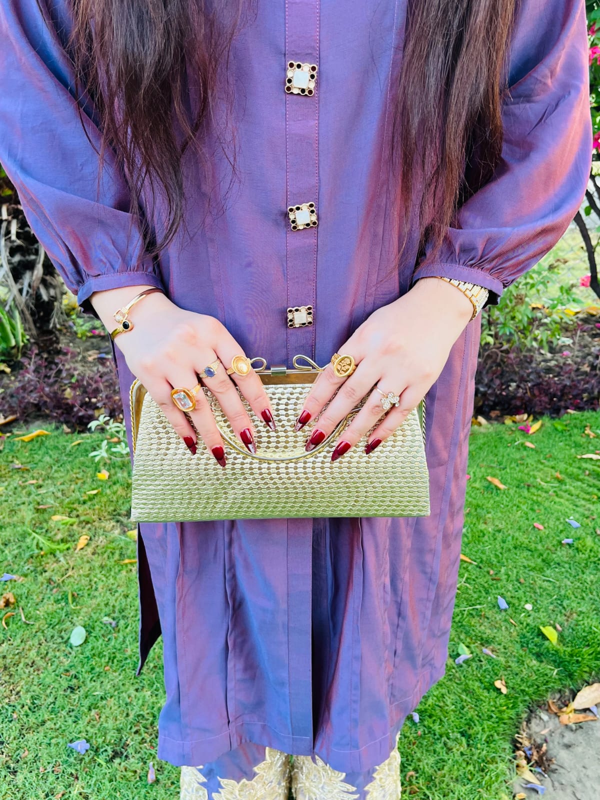 Girl wearing a purple outfit with gold buttons, holding a gold clutch against a green outdoor background